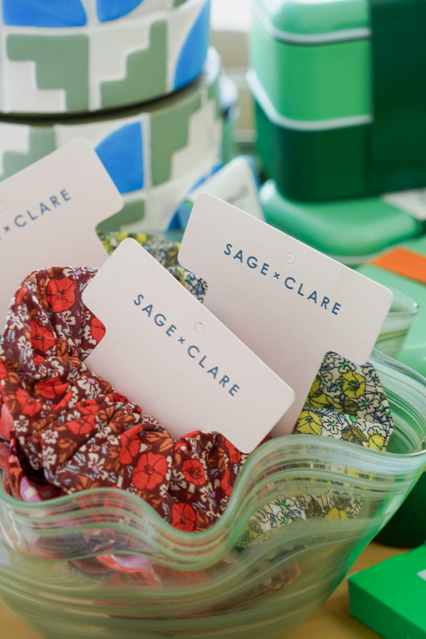 Floral Sage + Clare scrunchies displayed in a green, wavy glass bowl, with colorful boxes and patterns in the background.
